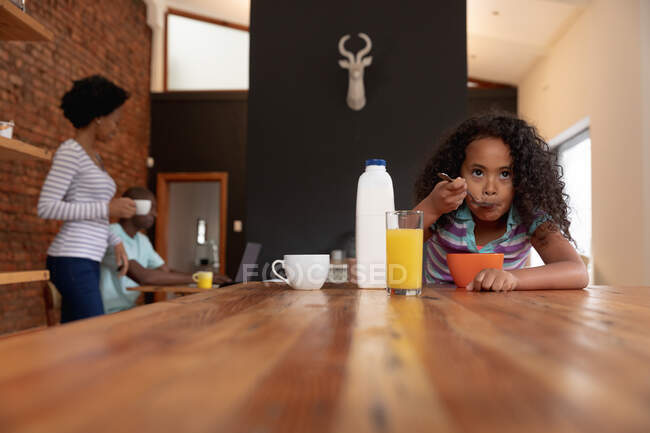 Front view of a young African American girl at home in the kitchen, sitting at a table eating breakfast cereal, her father sitting using a laptop computer and her mother standing beside him in the background — Stock Photo