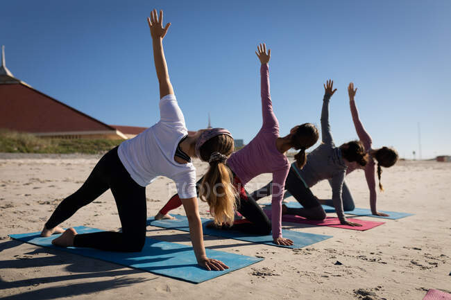 Вид сзади на многонациональную группу подруг, занимающихся спортом на пляже в солнечный день, практикующих йогу, растянувшихся в позиции йоги. — стоковое фото