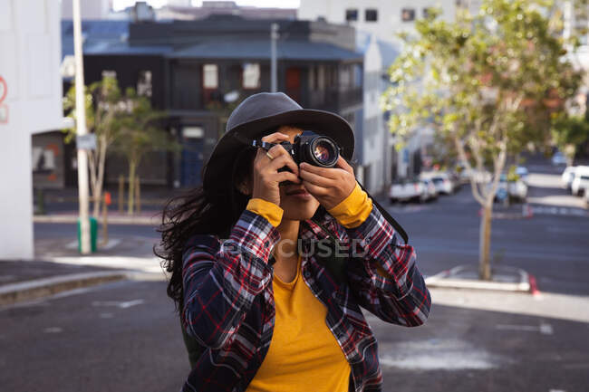 Vista frontal de una mujer de raza mixta con el pelo largo y oscuro por las calles de la ciudad durante el día, usando una cámara digital, usando un sombrero y camisa de cuadros y caminando por una calle de la ciudad con edificios en el fondo. - foto de stock