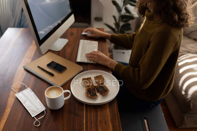 No meio de uma seção de uma mulher caucasiana passando tempo em casa, sentada ao lado de sua mesa e trabalhando usando seu computador, com um lanche e café colocados ao lado dela. Distanciamento social e auto-isolamento em quarentena . — Fotografia de Stock