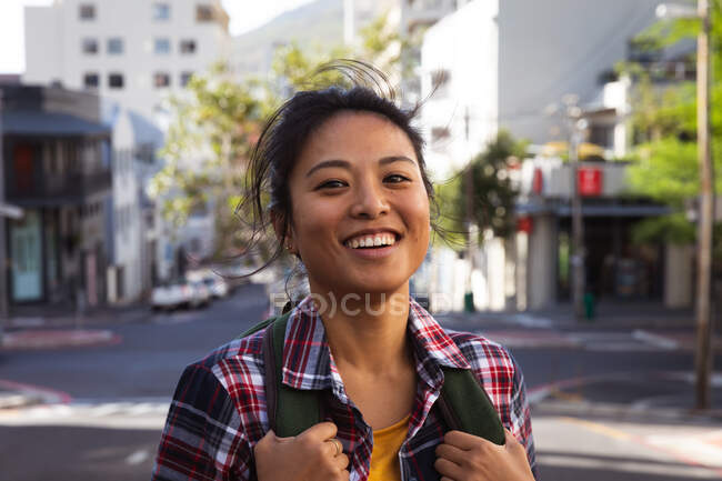 Porträt einer fröhlichen Mischlingshündin mit langen dunklen Haaren, die tagsüber auf den Straßen der Stadt unterwegs ist, einen Rucksack trägt, ein kariertes Hemd trägt und mit Gebäuden im Hintergrund in die Kamera lächelt. — Stockfoto