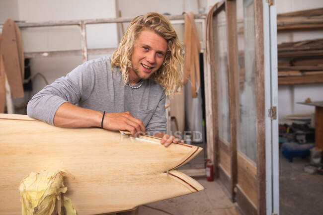 Caucasian male surfboard maker with long blond hair, in his studio, polishing a surfboard, looking at camera and smiling. — Stock Photo