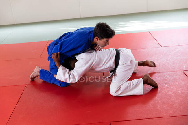 Side view of a mixed race male judo coach and teenage mixed race male judoka wearing blue and white judogi, practicing judo during a training in a gym. — Stock Photo
