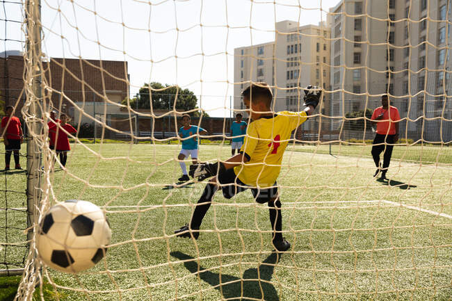 Передній вигляд Кавказького хлопчика, що дозволяє в голі грати в ворота з двома багатонаціональними командами футболістів, які грають в команді, під час футбольного матчу на футбольному полі. — стокове фото