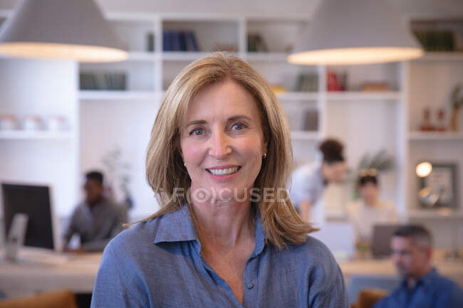 Retrato de uma empresária caucasiana feliz trabalhando em um escritório moderno, olhando para a câmera e sorrindo, com seus colegas trabalhando em segundo plano — Fotografia de Stock