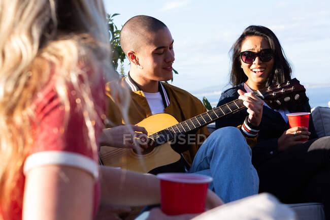 Vista frontal de un grupo multiétnico de amigos pasando el rato en una terraza en un día soleado, sonriendo, uno de ellos tocando la guitarra - foto de stock