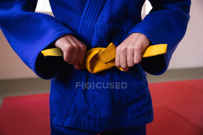 Vue de face section médiane de judoka debout sur des tapis de gym, attachant la ceinture jaune de judogi bleu. — Photo de stock