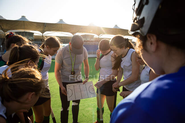 Side view of a group of female Caucasian field hockey players preparing before a game, huddling around their Caucasian male coach, on a hockey pitch, who gives them game plan, on a sunny day — Stock Photo