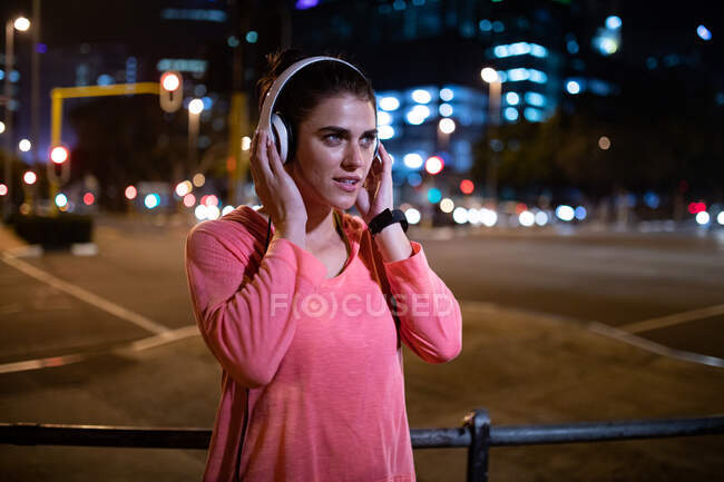 Vista frontal de uma mulher caucasiana apta com cabelos longos e escuros vestindo roupas esportivas exercitando-se ao ar livre na cidade à noite, fazendo uma pausa de seu treino em pé com fones de ouvido com edifícios urbanos no fundo . — Fotografia de Stock