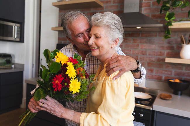 Ein älteres kaukasisches Ehepaar genießt seinen Ruhestand, steht an einem sonnigen Tag in der Küche, der Mann überreicht der Frau einen Blumenstrauß und küsst sie auf die Wange, zu Hause isoliert während der Coronavirus-Covid19 Pandemie — Stockfoto