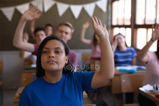 Vista frontal de una adolescente de raza mixta sentada en un escritorio en un aula y levantando las manos, sus compañeros de clase también levantando las manos en el fondo - foto de stock