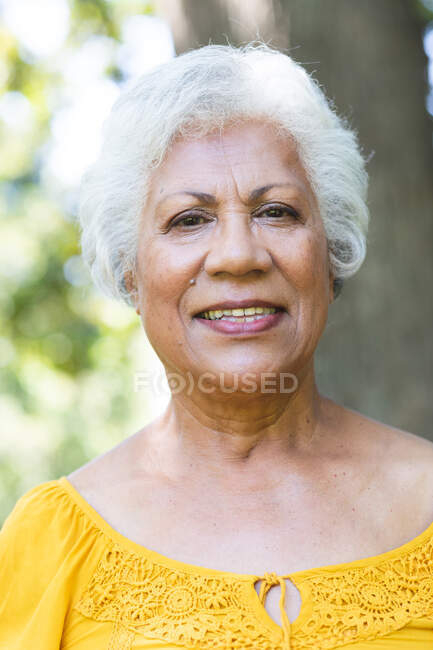 Retrato de perto de uma mulher afro-americana sênior atraente com cabelo branco curto desfrutando de sua aposentadoria em um jardim ao sol, olhando para câmera e sorrindo, auto-isolante durante a pandemia de coronavírus covid19 — Fotografia de Stock