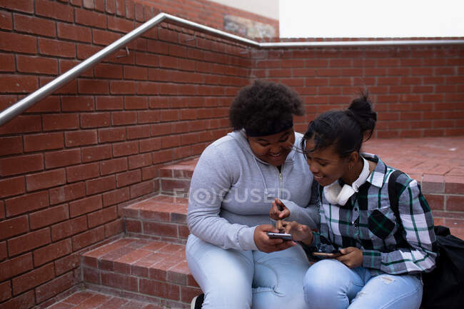 Seitenansicht von zwei afroamerikanischen Schülerinnen im Teenageralter, die ein Smartphone benutzen und auf Stufen auf dem Schulgelände sitzen — Stockfoto
