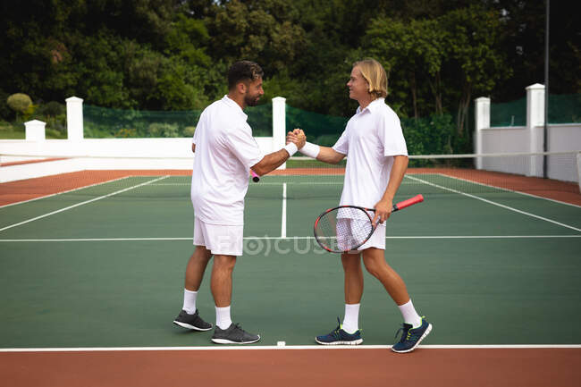 Un caucásico y un hombre de raza mixta que usan tenis blanco pasando tiempo juntos en una cancha, jugando al tenis en un día soleado, dándose la mano, uno de ellos sosteniendo una raqueta de tenis - foto de stock