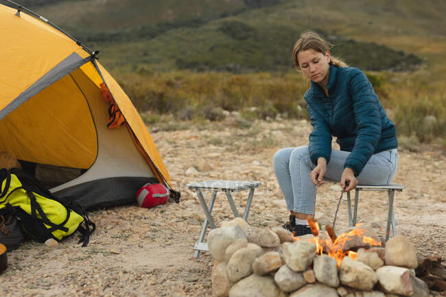 Vue latérale d'une femme caucasienne qui passe un bon moment en voyage à la montagne, assise près d'un feu de camp, cuisinant une saucisse sur un bâton — Photo de stock