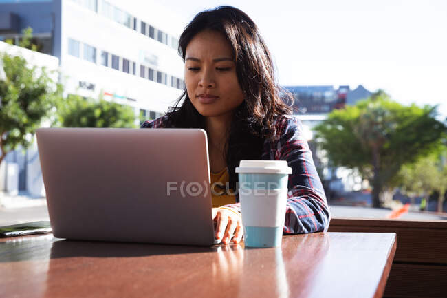 Vue de face d'une femme métisse aux longs cheveux foncés assise à une table dans un café pendant la journée, travaillant sur un ordinateur portable avec des bâtiments en arrière-plan. — Photo de stock