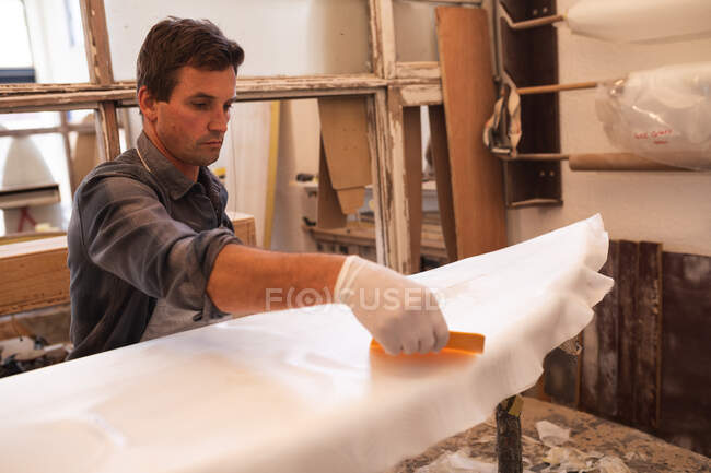 Homem caucasiano fabricante de pranchas de surf trabalhando em seu estúdio, preparando uma prancha de madeira coberta com um pedaço de pano branco para polir e pintar . — Fotografia de Stock