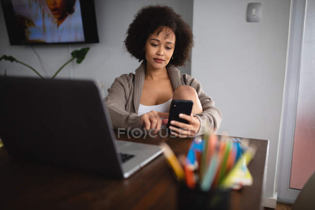 Vista frontal de cerca de una mujer de raza mixta sentada en una mesa en casa usando un teléfono inteligente, una computadora portátil y bolígrafos en la mesa frente a ella - foto de stock