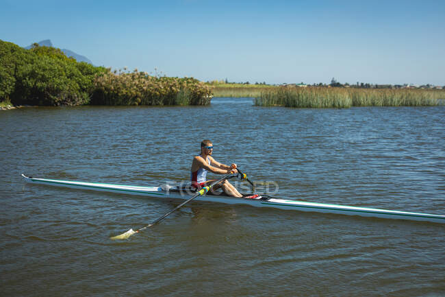 Seitenansicht eines kaukasischen männlichen Ruderers, der auf dem Fluss rudert, Ruder hält und an einem sonnigen Tag in einem Ruderboot sitzt — Stockfoto
