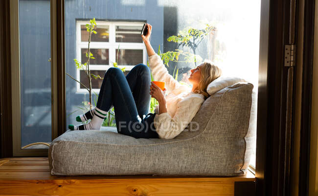 Seitenansicht einer kaukasischen Frau, die an einem sonnigen Tag in ihrem Wohnzimmer vor einem Fenster sitzt, ein Selfie macht und einen Becher hält — Stockfoto