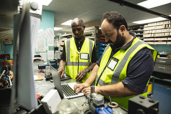 Kaukasische und afroamerikanische männliche Fabrikarbeiter mit Warnweste stehen an einer Werkbank und benutzen einen Laptop. Arbeiter in der Industrie in einer Fabrik für hydraulische Anlagen. — Stockfoto