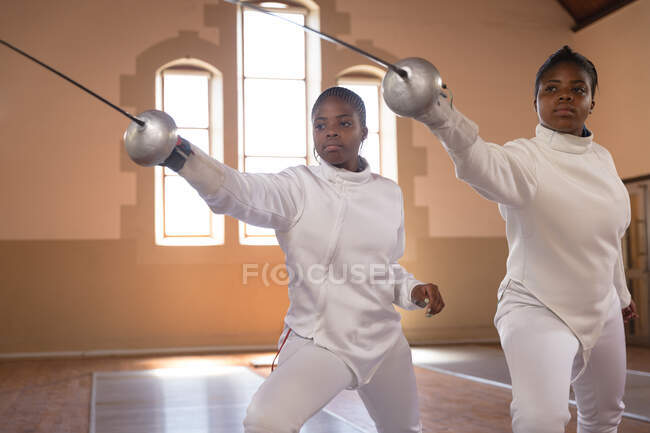 Des sportives afro-américaines portant des tenues d'escrime protectrices lors d'une séance d'entraînement à l'escrime, tenant des épees et se jetant à l'unisson. escrimeurs entraînement dans une salle de gym. — Photo de stock