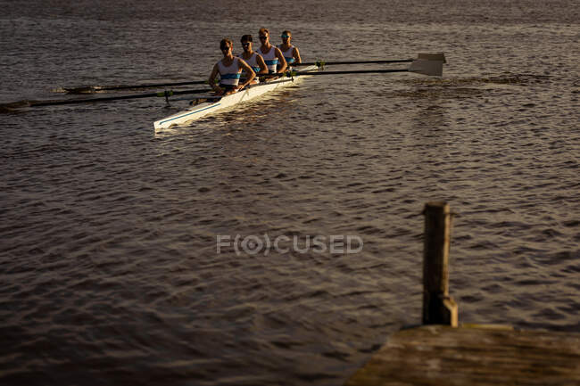 Vista frontale di una squadra di canottaggio di quattro uomini caucasici che si allenano e remano sul fiume, seduti su una barca a remi che regge remi — Foto stock