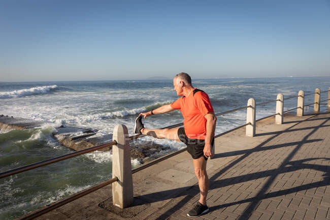 Vista lateral de un hombre mayor caucásico maduro haciendo ejercicio en un paseo marítimo en un día soleado con cielo azul, estirándose con una pierna en una balaustrada - foto de stock