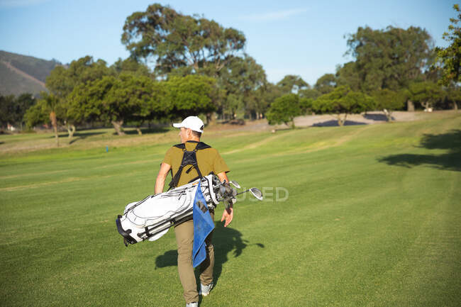Вид сзади на кавказца на поле для гольфа в солнечный день с голубым небом, гуляющего и несущего сумку для гольфа — стоковое фото