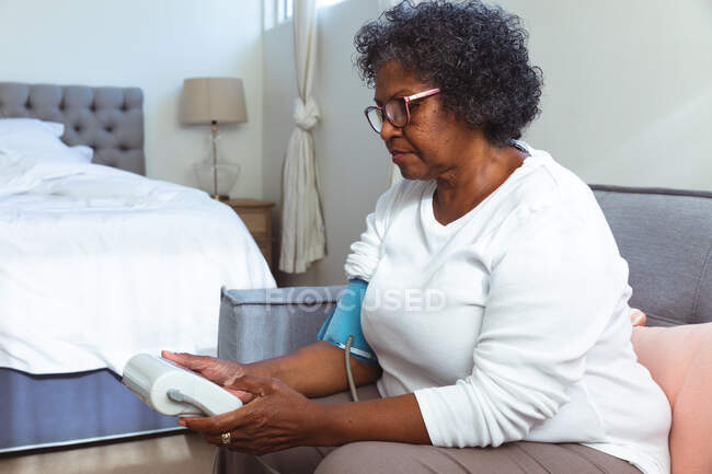 Mujer de raza mixta mayor pasando tiempo en casa, sentada en una silla y tomando su presión arterial con un tonómetro, distanciamiento social y autoaislamiento en cuarentena - foto de stock