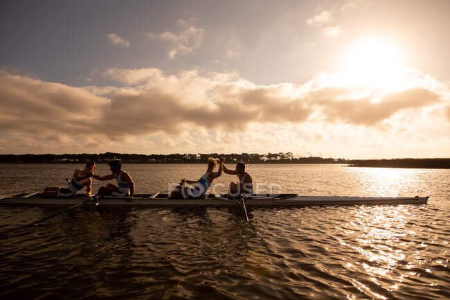 Side view of a rowing team of four Caucasian men training and rowing on the river, sitting in a rowing boat, holding their hands, celebrating and enjoying the training — Stock Photo