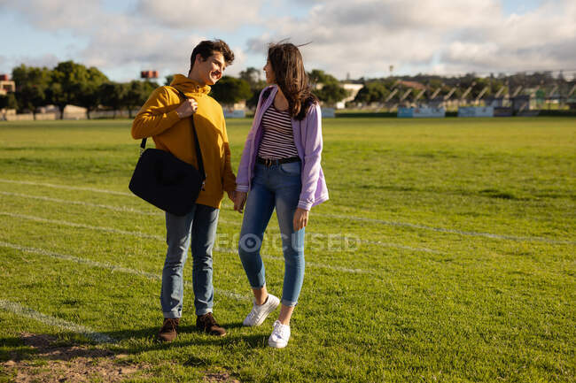 Вид спереди кавказской девочки-подростка и мальчика, держащихся за руки и улыбающихся друг другу, стоящих на школьном игровом поле — стоковое фото