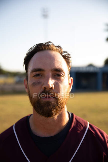 Retrato de un jugador de béisbol caucásico, vistiendo un uniforme de equipo, parado en un campo de béisbol, mirando una cámara - foto de stock