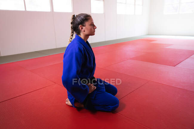 Vista laterale di una judoka caucasica adolescente vestita con judogi blu, inginocchiata sui tappetini in palestra prima dell'allenamento con il judo. — Foto stock