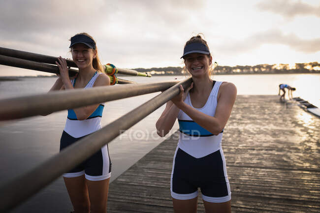 Передня сторона дивиться на двох кавказьких жінок з веслувального загону з веслувальними веслами на плечах і йде по пристані на річці на світанку, посміхаючись до камери — стокове фото
