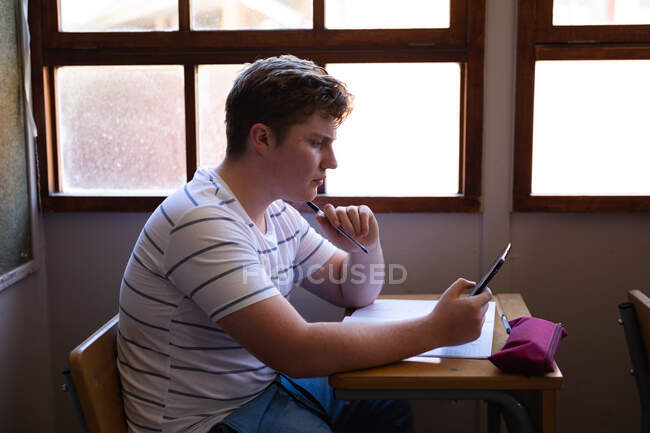Seitenansicht eines kaukasischen Teenagers, der mit einem Smartphone am Schreibtisch im Unterricht sitzt und sich konzentriert — Stockfoto
