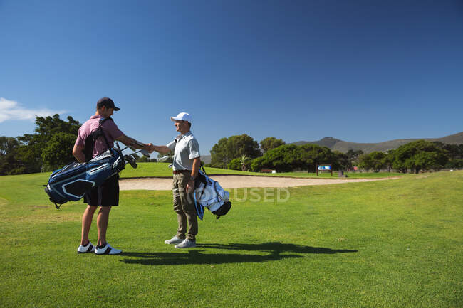 Side view of two Caucasian men at a golf course on a sunny day with blue sky, shaking hands while carrying golf bag — Stock Photo