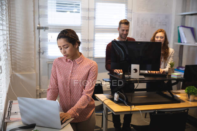 Eine kreative Unternehmerin mit gemischter Rasse, die in einem lässigen modernen Büro arbeitet, am Schreibtisch steht und Laptop benutzt, während eine männliche und weibliche Kollegin an einem Stehpult im Hintergrund arbeiten. — Stockfoto