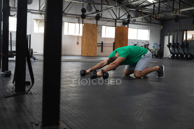 Vue latérale d'un homme athlétique caucasien portant des vêtements de sport cross-training dans une salle de gym, musculation avec haltères, à genoux, penché vers l'avant et s'étirant, tenant un haltère dans chaque main — Photo de stock