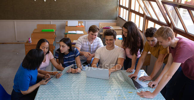 Vista de ángulo alto de un grupo multiétnico de alumnos adolescentes sentados y parados alrededor de una mesa en un salón de clases mirando juntos computadoras portátiles y tabletas y hablando en tiempo de descanso - foto de stock
