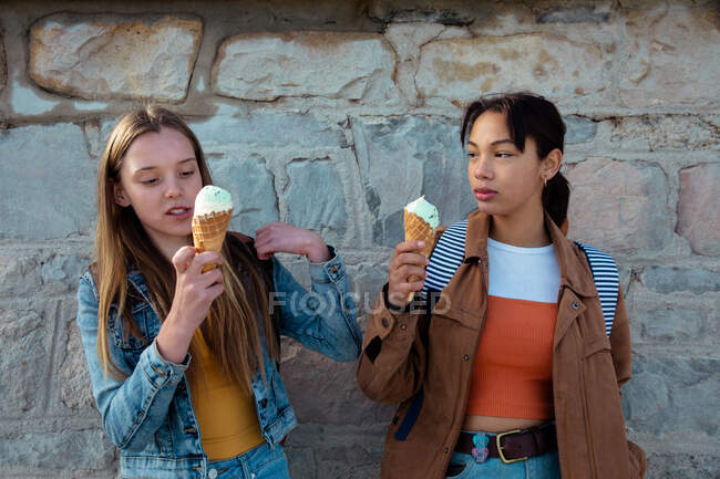 Vista frontal de um caucasiano e uma mista meninas de raça desfrutando de tempo pendurado juntos em um dia ensolarado, comer sorvete, de pé junto à parede . — Fotografia de Stock