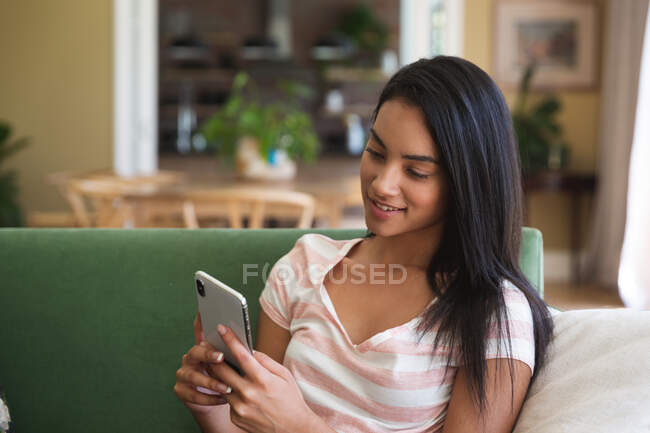 Mixed race woman spending time at home self isolating and social distancing in quarantine lockdown during coronavirus covid 19 epidemic, sitting on a sofa using smartphone in sitting room. — Stock Photo