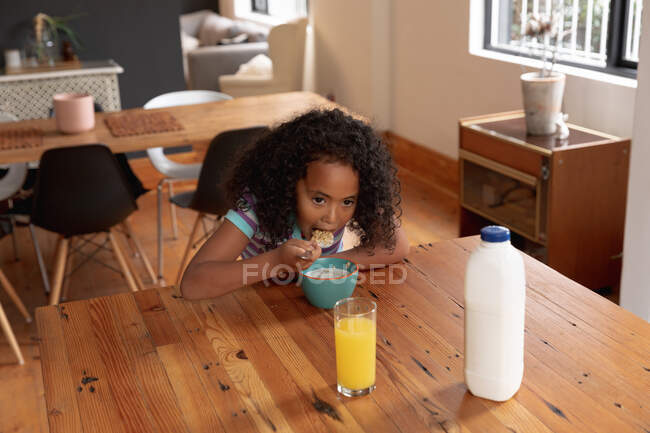 Висококутний вид на молоду афроамериканську дівчину вдома на кухні, сидить за столом і їсть сніданок пластівці, склянку апельсинового соку і пляшку молока на столі перед нею. — стокове фото