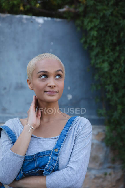 Mixed race alternative woman with short blonde hair out and about in the city on a sunny day, standing by a wall and looking away. Urban independent woman on the go. — Stock Photo
