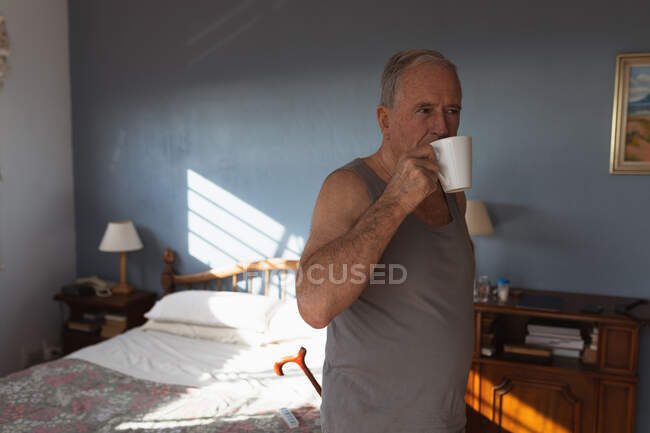 Вид сбоку на пожилого кавказца, расслабляющегося дома в своей спальне, стоящего и пьющего чашку кофе после утреннего пробуждения в солнечный день — стоковое фото