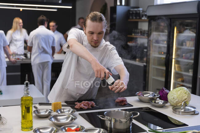 Cuoco maschio caucasico che aggiunge sale all'acqua bollente, con altri chef che cucinano sullo sfondo. Classe di cucina in una cucina ristorante. — Foto stock