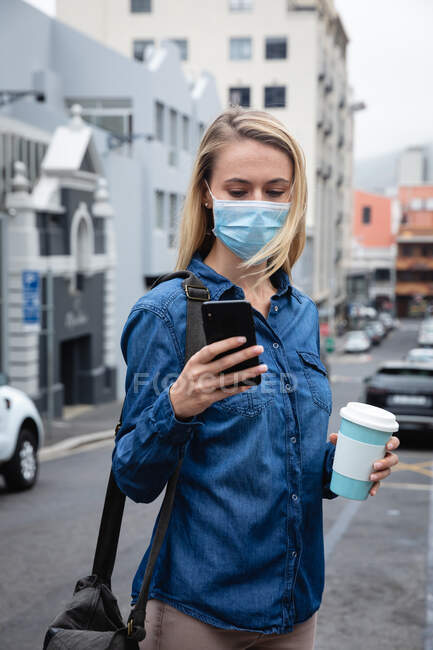 Vista frontal de cerca de una mujer caucásica con máscara facial contra la contaminación del aire y covid19 coronavirus, caminando por las calles de la ciudad, usando su teléfono inteligente y sosteniendo una taza de café para llevar. - foto de stock