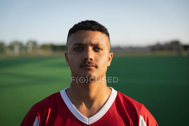 Portrait close up of a confident mixed race male field hockey player, wearing a red team strip, standing on a hockey pitch looking to camera on a sunny day — Stock Photo