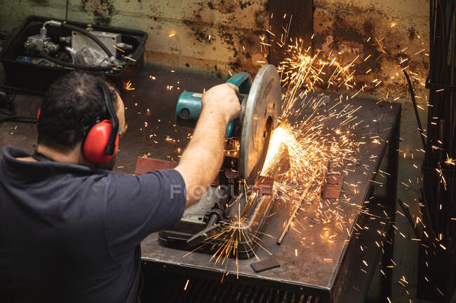 Operaio caucasico di fabbrica con grembiule scuro e occhiali di sicurezza, in piedi su un banco da lavoro e tagliare un metallo in un'officina. — Foto stock