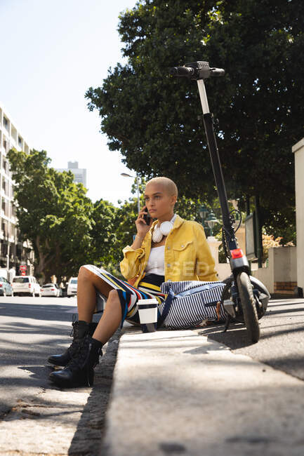 Mujer alternativa de raza mixta con pelo corto y rubio en la ciudad en un día soleado, sentada en la acera con smartphone, e-scooter y un café a su lado. Nómada digital urbano en movimiento. - foto de stock